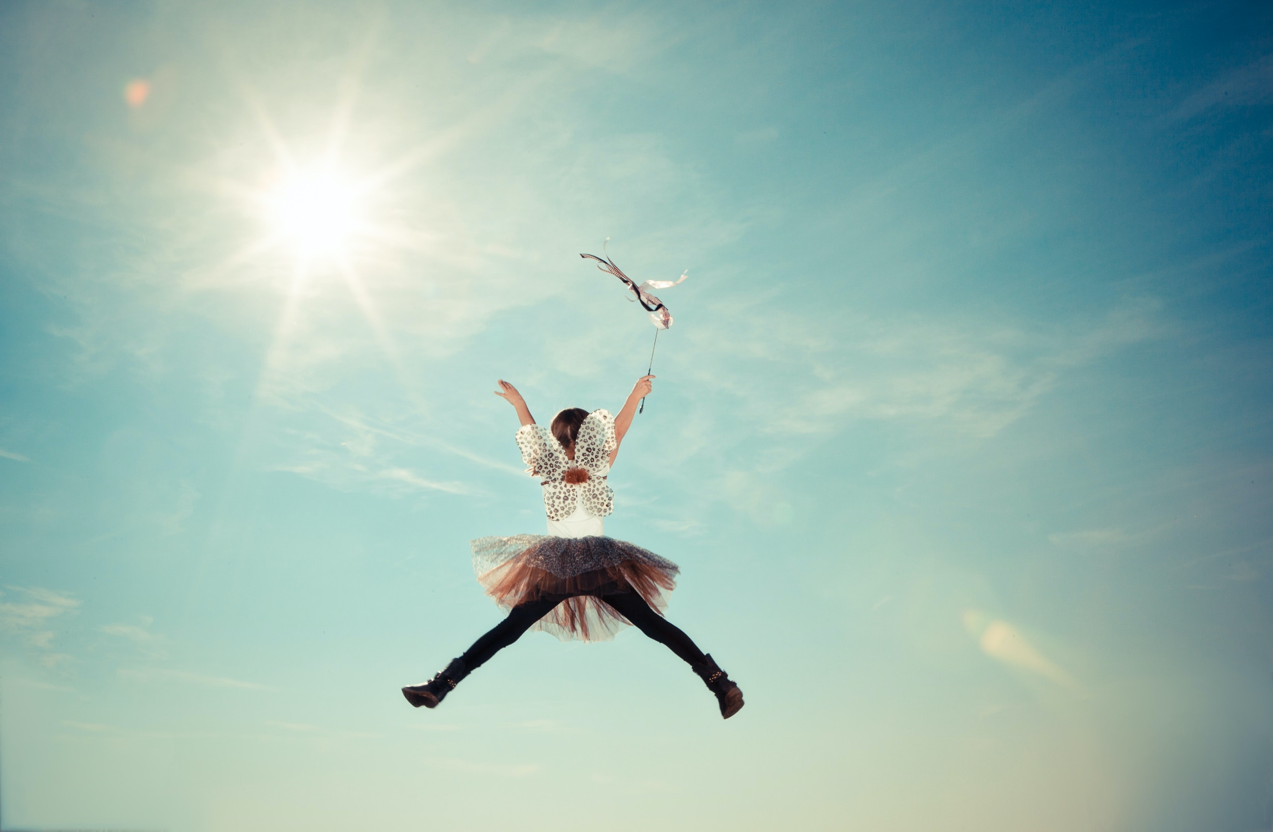 Weibliche Person springt in die Luft vor einem blauen Himmel und Sonne. Sie ist von hinten zu sehen und spreizt die arme und Bein von sich um eine X zu formen.
