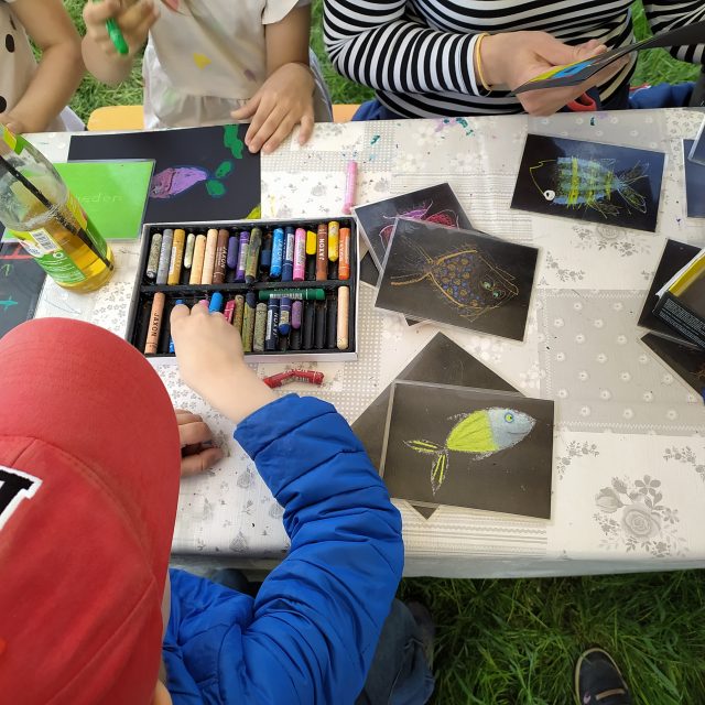 Kinder an einem Basteltisch, die Fische und Gefühle malen, im Vordergrund ein Kind mit rotem Capy und blauer Jacke.