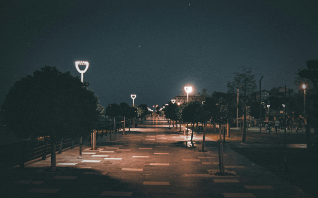 Eine ruhige, breite Promenade bei Nacht, beleuchtet von modernen Straßenlaternen, die in regelmäßigen Abständen entlang des Weges stehen. Auf beiden Seiten der Promenade sind kleine Bäume und Sträucher zu sehen, und im Hintergrund sind schemenhaft Gebäude und weitere Lichter erkennbar. Die Atmosphäre wirkt friedlich und still.