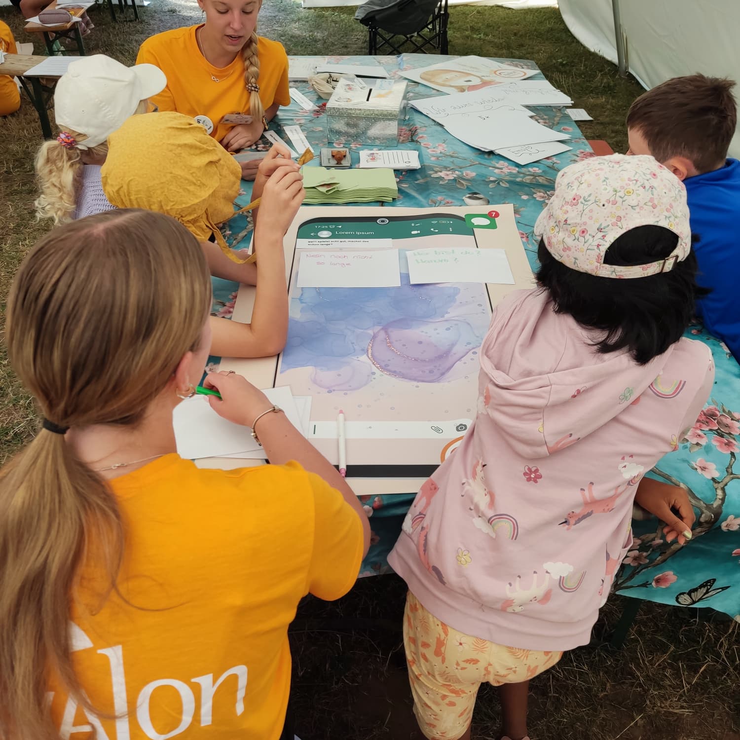 Eine Gruppe von Kindern und erwachsenen um einen Tisch. Sie besprechen Regeln zum sicheren Umgang im Chat am Handy. Die Erwachsenen tragen Apricotfarbene T-shirts mit dem Logo der Fachberatungsstelle in weiß.